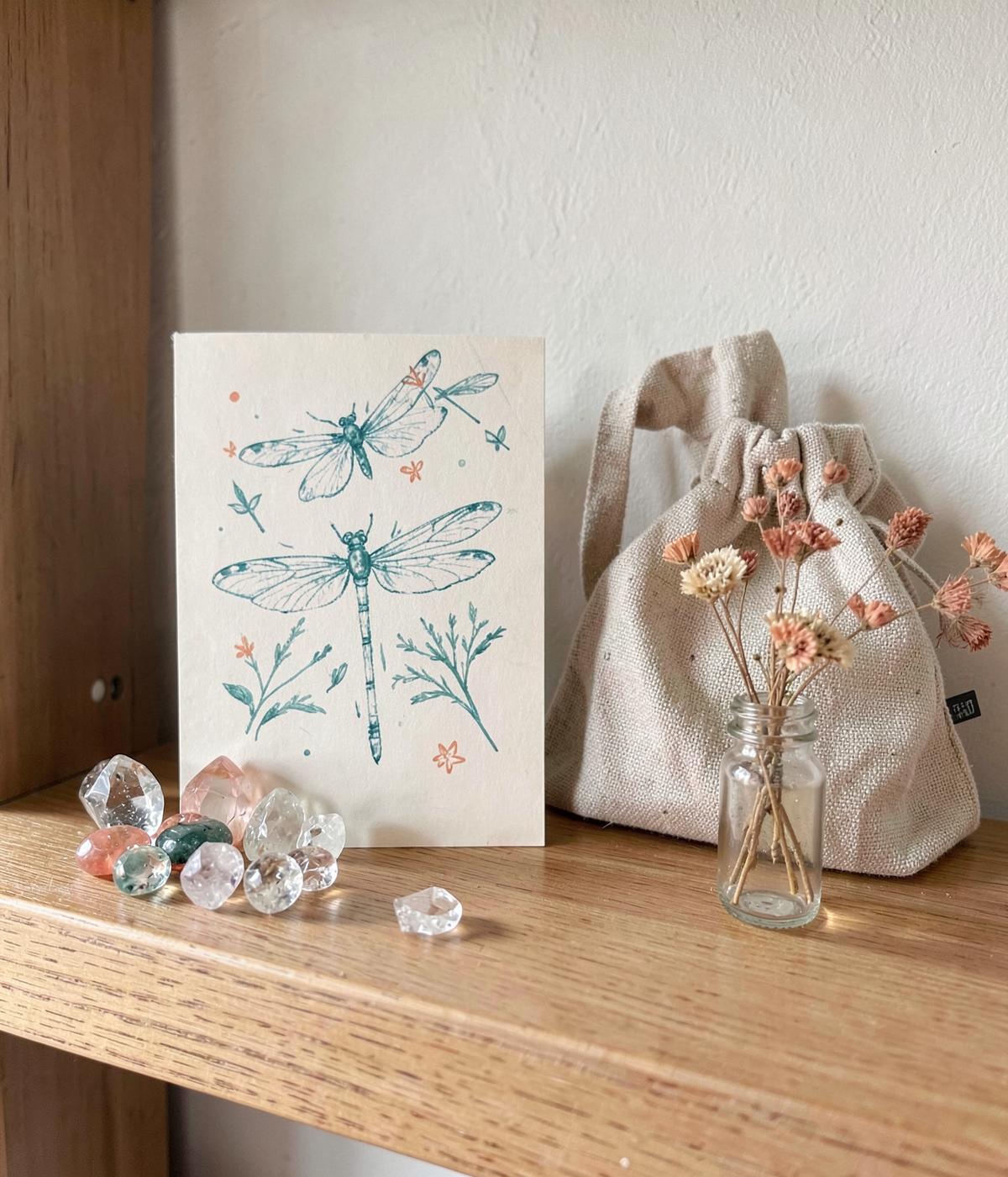 Hand-illustrated dragonfly card with crystals and a linen gift pouch on a wooden shelf